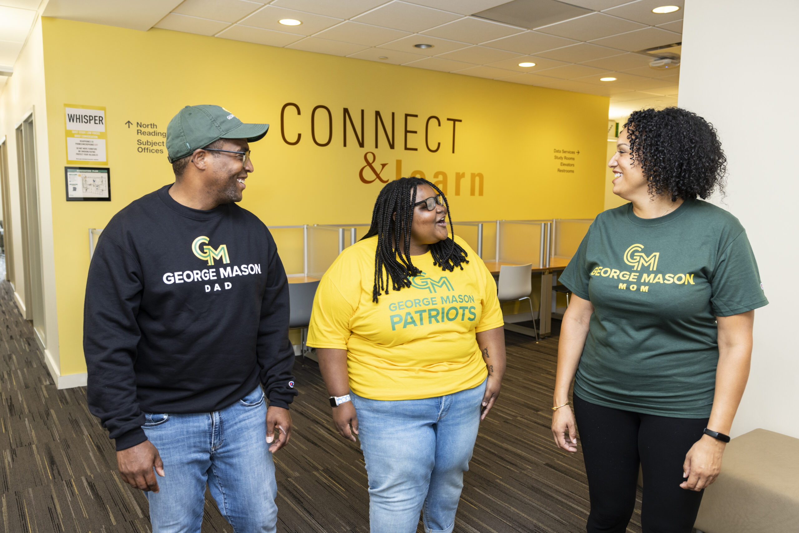 Students wear new Mason Gear. Photo by: Ron Aira/Creative Services/ George Mason University Three people smile at each other in a hallway.