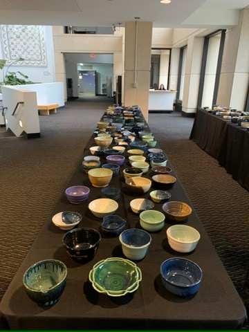 A long table covered in a black cloth is filled with rows of colorful handmade ceramic bowls in various shapes and glazes, arranged in a hallway or lobby with carpeted floor, tall windows, and additional tables of bowls visible in the background.