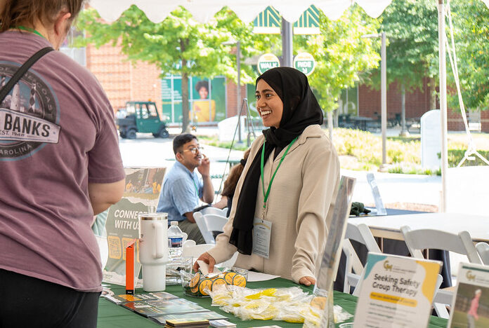 Three students walking together down a street lined with shops and greenery, one wearing a 'Patriot Pride' shirt.