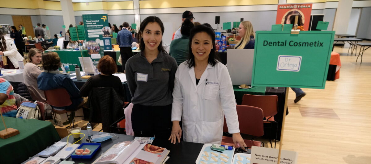 Two people standing in front of a table at the Health and Fitness Expo, with a number of flyers spread out on the table.