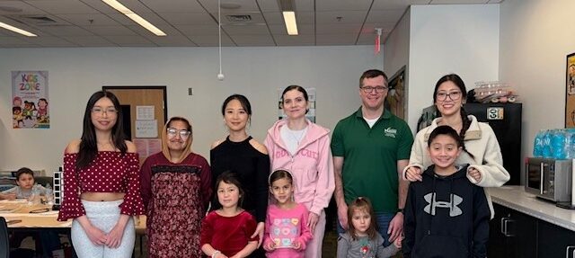 A group of student parents, staff members, and children pose together for a photo at a CSS Student Parent and Caregiver Meetup. A "Kids Zone" sign is visible in the background.