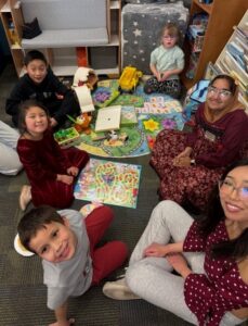 Children and two adults sit on the floor of a kids' area, playing board games together at a CSS Student Parent and Caregiver Meetup.