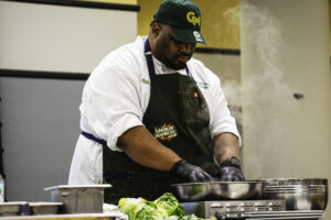 A chef wearing a white coat, green George Mason cap, and dark apron works at a stainless steel cooking station, with steam rising and fresh vegetables visible on the counter.