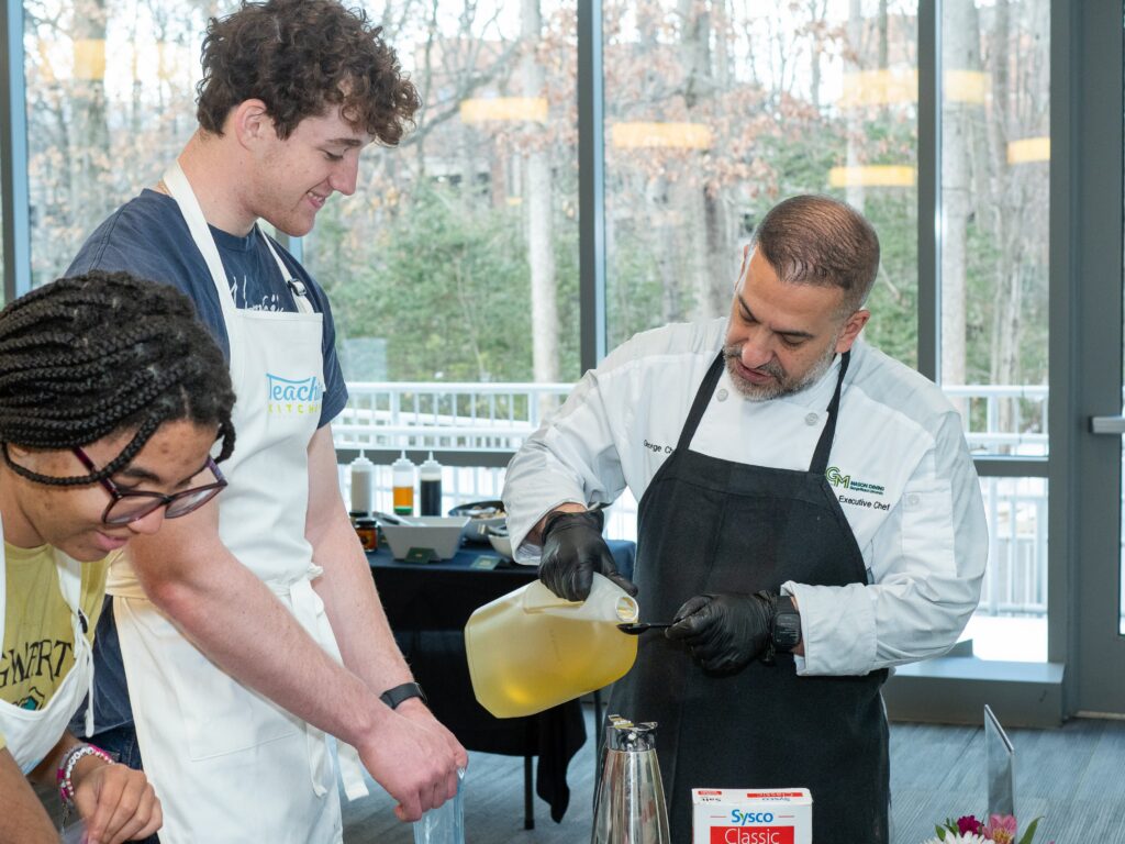 Campus Executive Chef George Charbel demonstrates a cooking technique for students during a Teaching Kitchen session, as participants measure ingredients at a hands-on station.