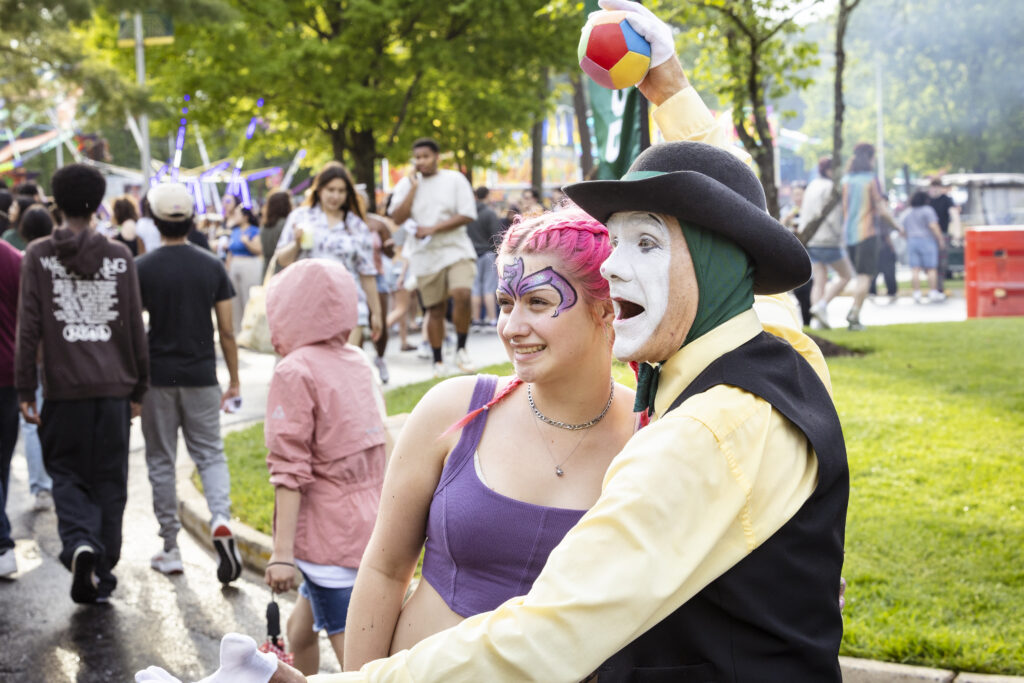 A student with pink hair and butterfly face paint smiles next to a mime performer in a black hat and white face paint at an outdoor campus festival.