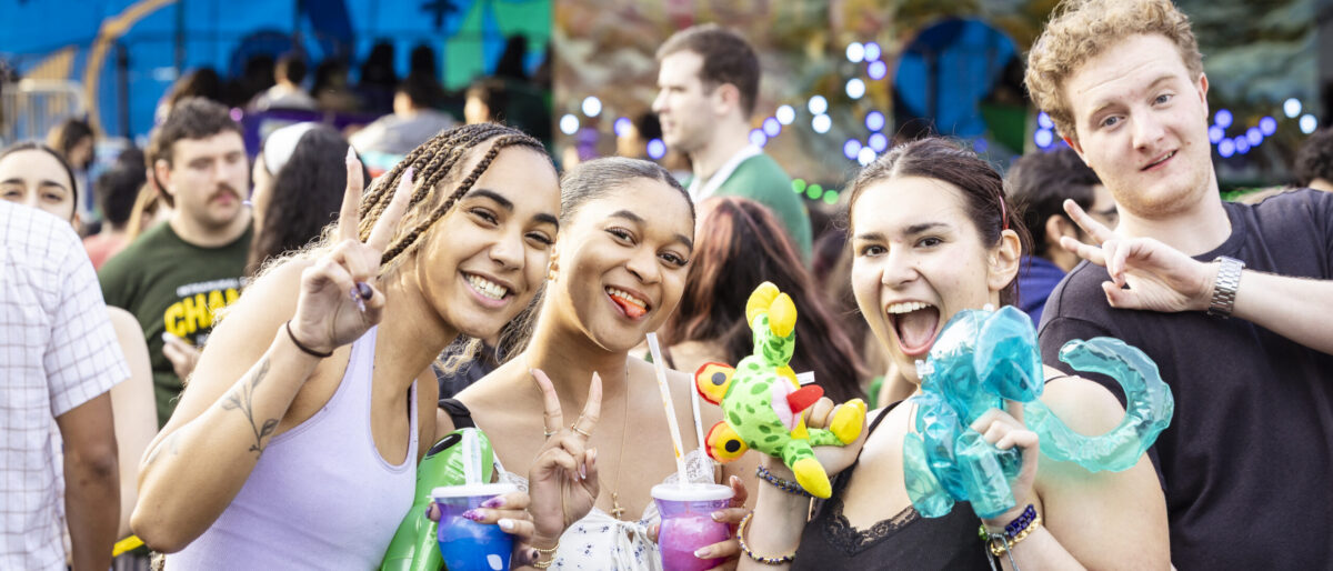 Four students smile and hold colorful inflatable carnival prizes and drinks in front of a lit carnival backdrop.