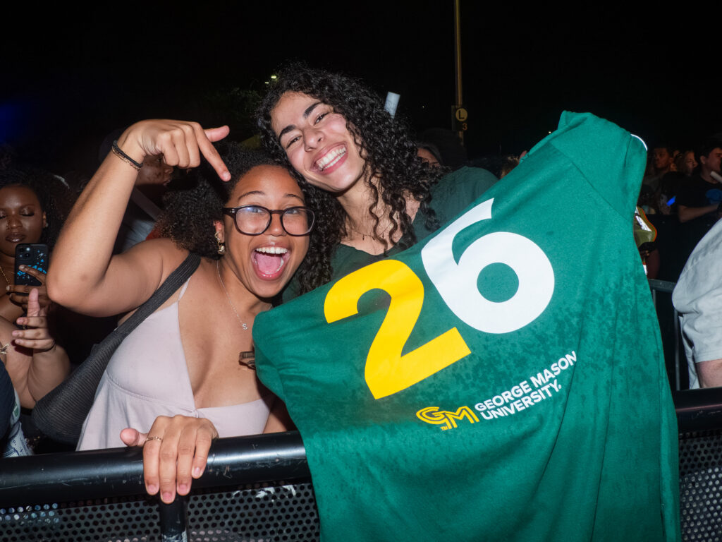 Two students smile and hold up a green George Mason University Class of 2026 shirt at a nighttime outdoor concert.