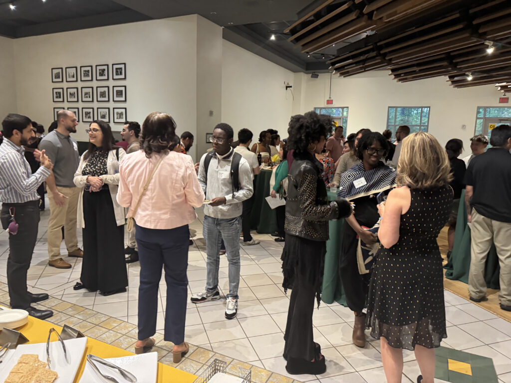 A group of people in professional attire engage in conversation at an indoor networking event. Several wear name tags. Additional attendees are visible in the background.
