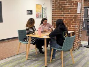 Three people sit around a small table in a campus building, looking at cards laid out on the table. Two cups and a water bottle are visible. One person, facing the camera, wears a hoodie with a Student Strengths Coach logo on the back.