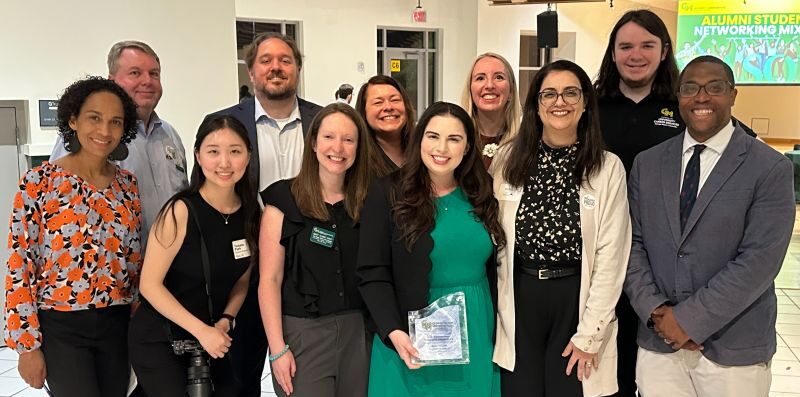 11 people stand smiling in front of a projected screen reading "Alumni Student Networking Mixer" at George Mason University. They wear name tags and professional attire.