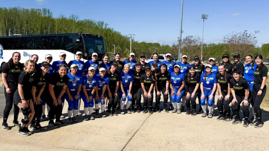Members of the George Mason softball team in black uniforms and the Saint Louis Billikens in blue uniforms pose together in two rows in front of a white charter bus outside the softball complex on a sunny spring afternoon.