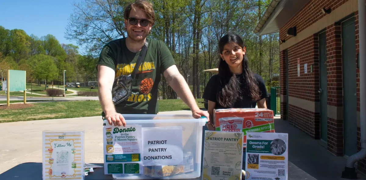 John Eichler and a female student stand behind an outdoor donation table displaying a clear plastic bin labeled "Patriot Pantry Donations," a case of Cup Noodles, Patriot Pantry informational signage, and a "Gowns for Grads" flyer. Trees and a brick building are visible in the background.