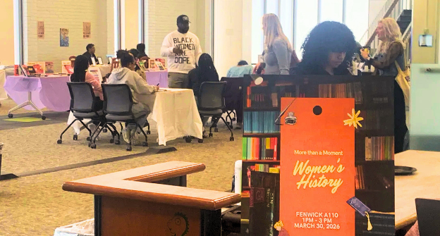 A wide shot of a library event space with students seated at tables and browsing books. A colorful orange event sign reading "More Than a Moment: Women's History Month, Fenwick A110, 1–3 p.m., March 30, 2026" stands in the foreground.