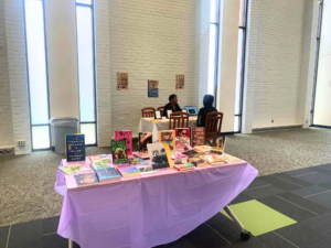 A purple-draped table covered with a wide variety of books in the foreground. Two people sit at a smaller table in the background against a white brick wall with small artworks displayed between windows.