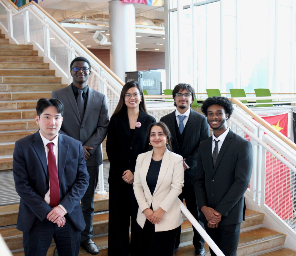 Six students dressed in formal business attire pose together on the stairs of the Johnson Center. International flags are visible hanging from the upper level in the background. Duong Thuy Nguyen, wearing a black outfit with a floral pin, stands in the middle of the back row surrounded by her Capstone teammates.