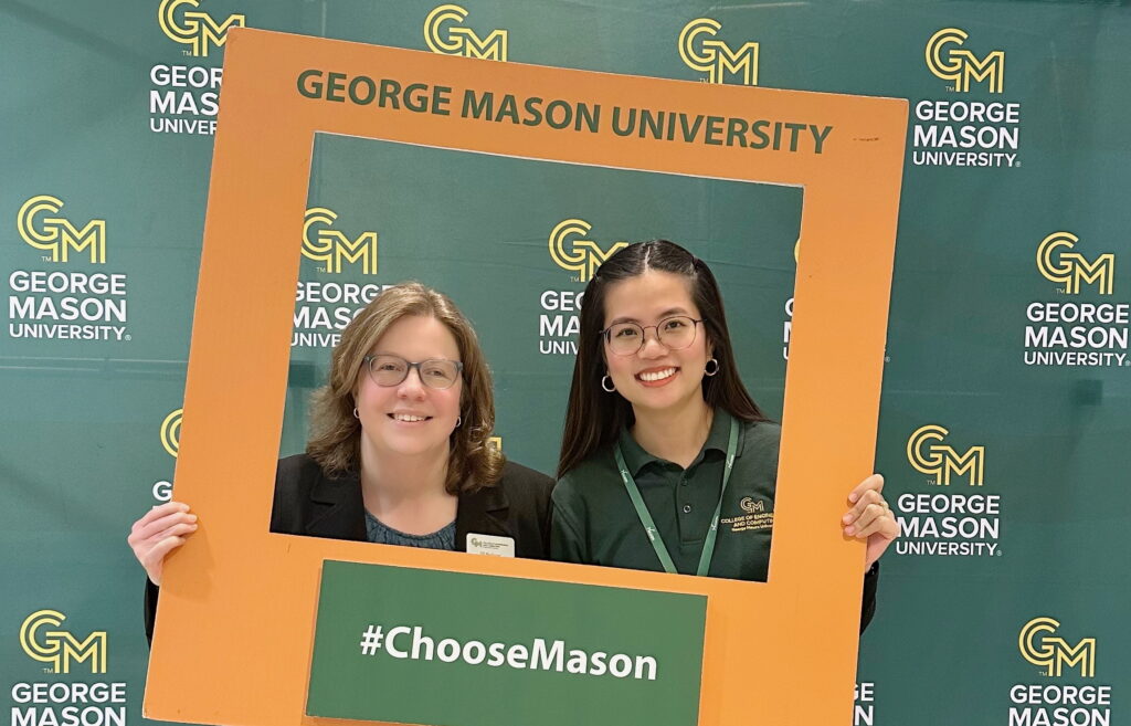 From left, Associate Dean Jill Nelson and Duong Thuy Nguyen smile while holding an oversized orange photo frame printed with "George Mason University" and "#ChooseMason" in front of a green George Mason University step-and-repeat banner. Nguyen is wearing a green College of Engineering and Computing polo and a George Mason lanyard.