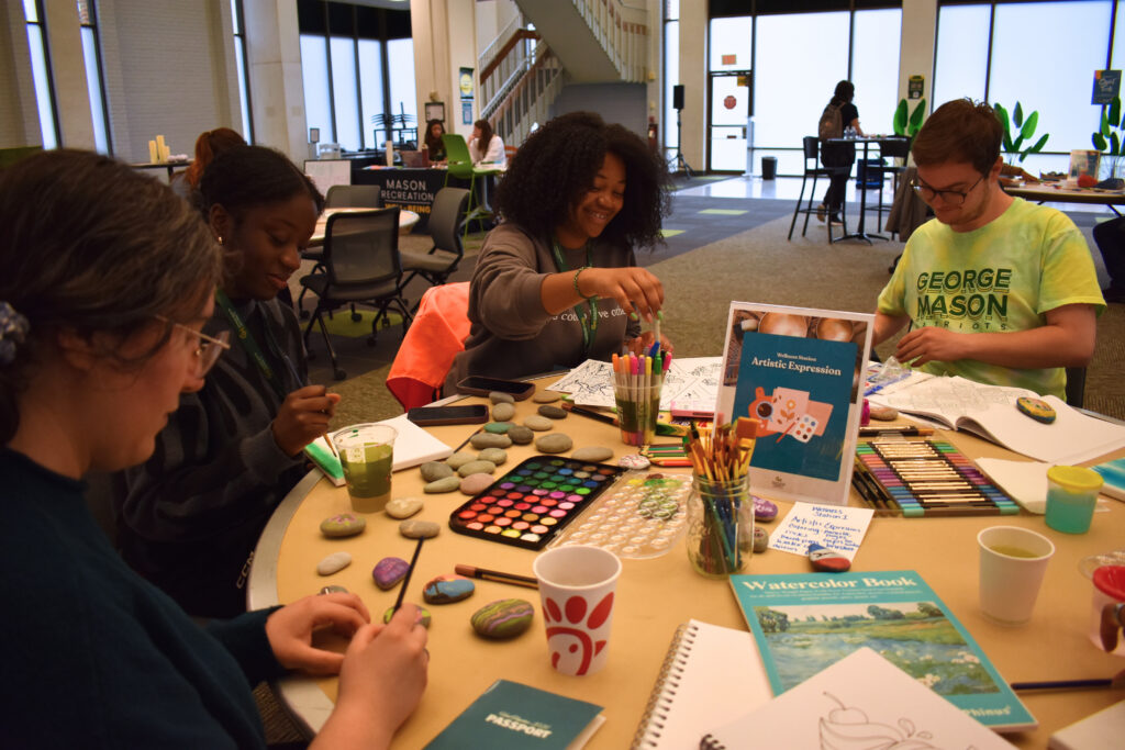 Four students sit around a round table covered with art supplies including painted rocks, watercolor palettes, brushes, markers, and watercolor books. A student in the center smiles while reaching across the table. A student on the right wears a green George Mason University t-shirt. A "Mason Recreation" sign is visible in the background.