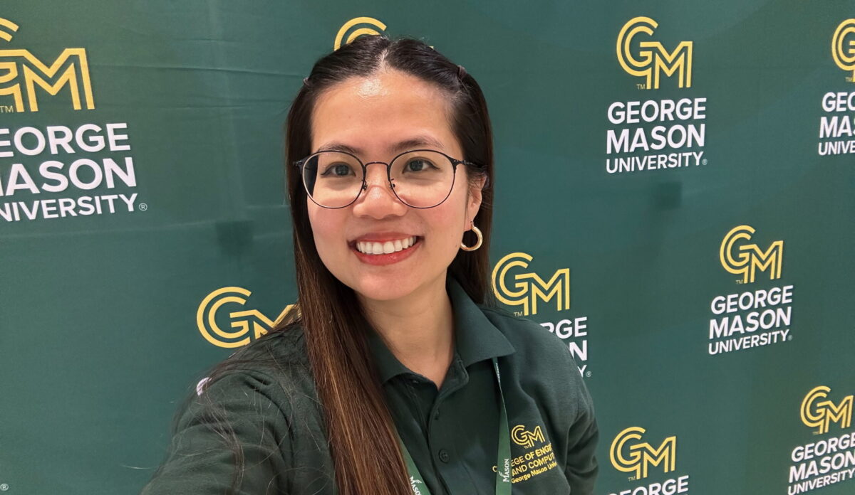 Duong Thuy Nguyen, wearing a green College of Engineering and Computing polo and a George Mason lanyard, smiles for a selfie in front of a green George Mason University step-and-repeat banner.