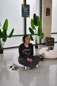 A student sits cross-legged on a round floor cushion, eyes closed, wearing over-ear headphones and glasses. A "Welcome to Quiet Tech" sign is visible on the wall behind her, flanked by large tropical plants. A second cushion with headphones rests nearby.