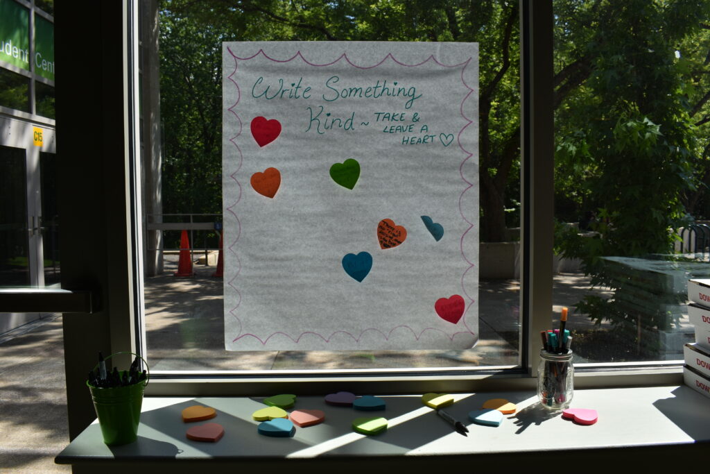 A handmade poster reading "Write Something Kind — Take and Leave a Heart" hangs in a sunlit window, decorated with colorful fabric hearts. Below it, a windowsill holds a small green pot, a jar of markers, scissors, and cutout paper hearts in yellow, pink, and teal.