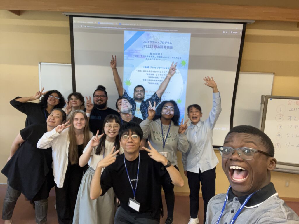 Simeon Omorodion laughs in the foreground of a group selfie with approximately ten classmates, all smiling and making peace signs. Behind them, a projected slide displays the title of a 2024 summer Japanese language presentation. A whiteboard with Japanese writing is visible to the right.