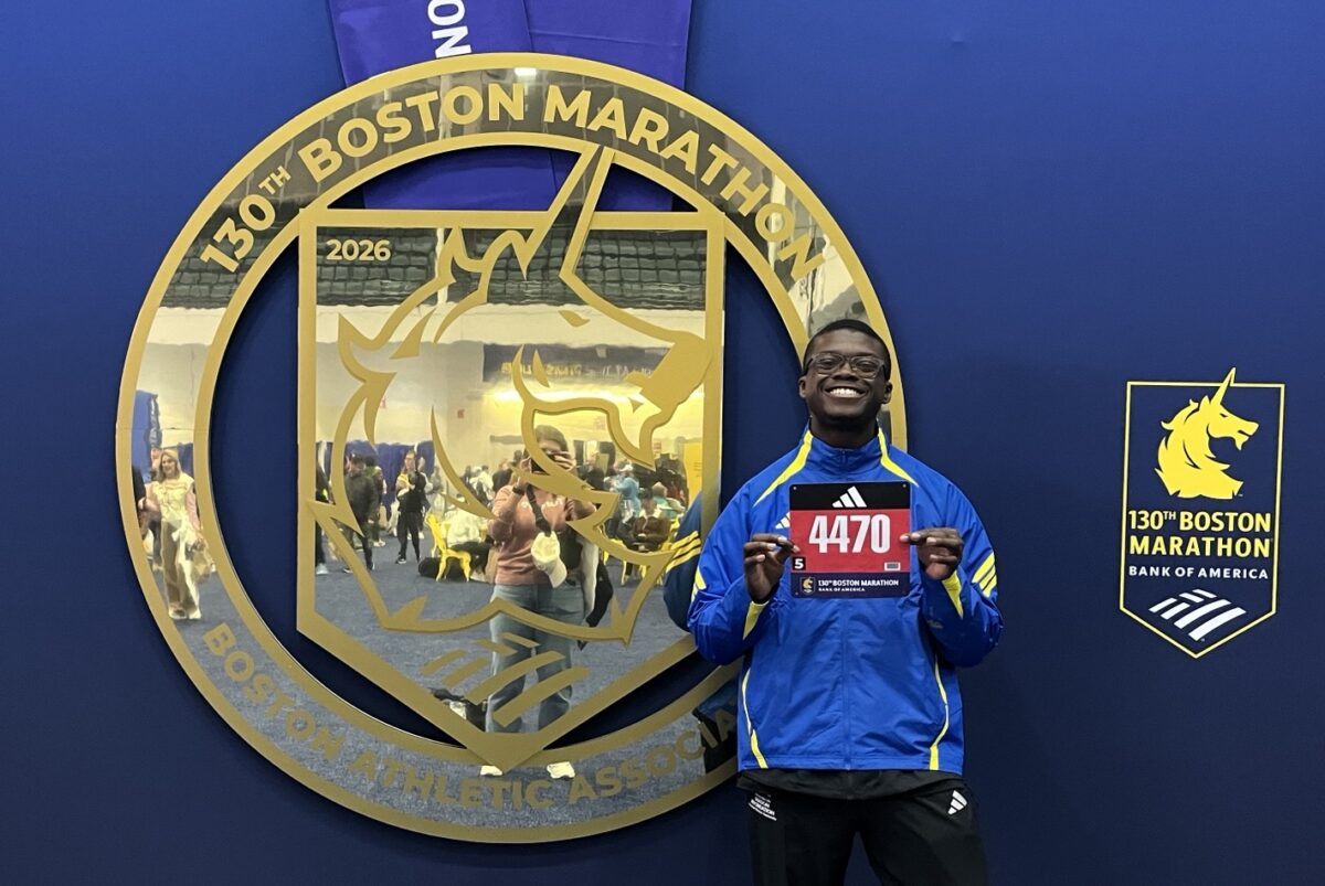 Simeon Omorodion smiles and holds up his race bib, number 4470, in front of a large decorative medal display for the 130th Boston Marathon. He is wearing a blue and yellow track jacket and black pants.