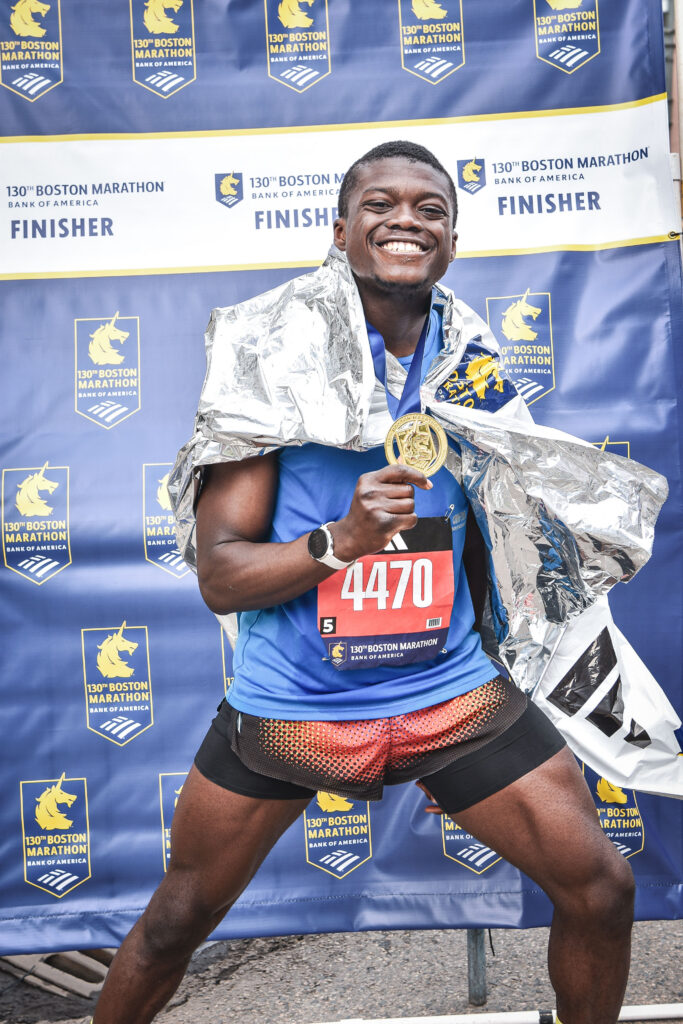 Simeon Omorodion stands in front of a 130th Boston Marathon finisher backdrop, smiling broadly and holding up his finisher's medal. He is wearing a blue race jersey with bib number 4470, red and black shorts, and a silver thermal blanket draped over his shoulders.