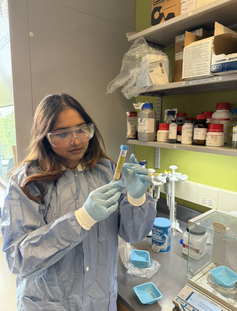Zarin Musarrat Niladree stands at a laboratory bench wearing a blue lab coat, safety goggles, and blue gloves, examining a test tube with a yellow substance. Shelves with chemical bottles and lab supplies are visible in the background.