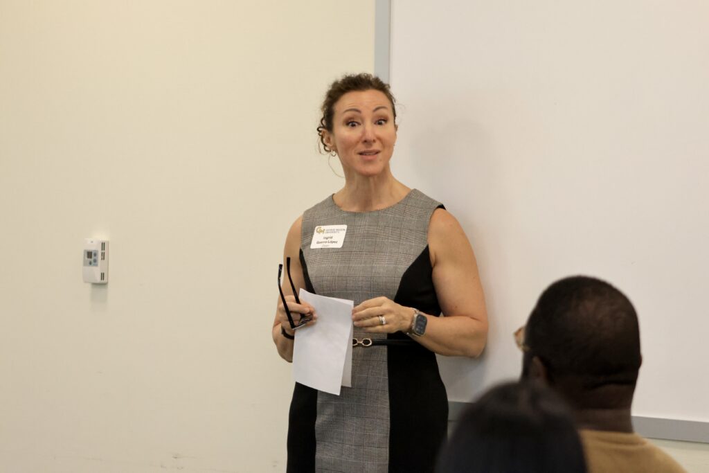 Ingrid Guerra-Lopez stands at the front of a classroom holding notes and glasses while addressing an audience. She wears a gray and black sleeveless dress and a George Mason University name tag.
