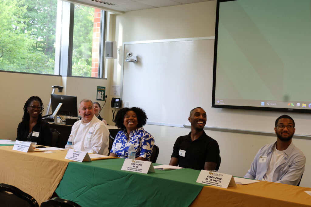Five panelists sit behind a table draped in green and gold, laughing and smiling. From left: Alicia Brown, Thomas Ammazzalorso, Tia Pierce, Luke Pierce, and Cedric Price. Name placards with George Mason University branding are visible in front of each panelist.